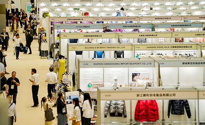 Trade fair floor at Mydome Osaka, rows of exhibitor booths with garment rails and buyers walking the aisles